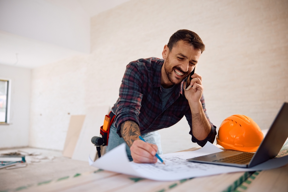 Happy,Construction,Worker,Communicating,On,Cell,Phone,While,Analyzing,Blueprints